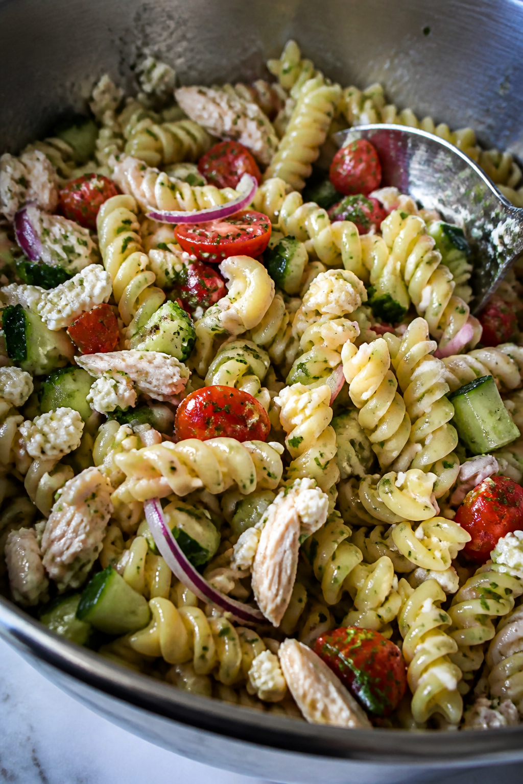 Close-up detail: Tossed chicken feta cucumber pasta salad mid-mix in a wide stainless-steel bowl, al
